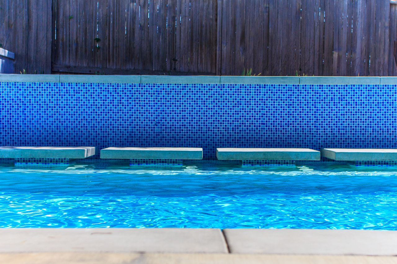 Swimming pool with blue tiled wall, stone stepping pads, and wooden fence.