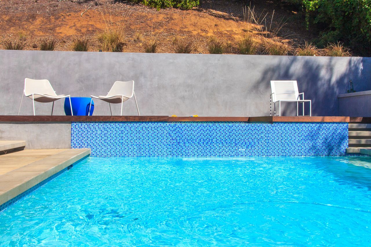 Swimming pool with blue tiles, wooden deck, white chairs, and a grey wall.
