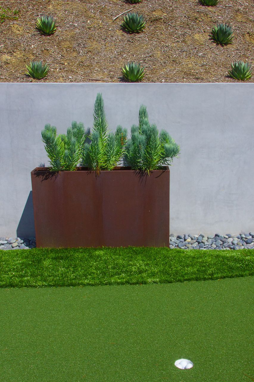 A rectangular brown planter with green plants against a gray wall, bordered by rocks and artificial turf.