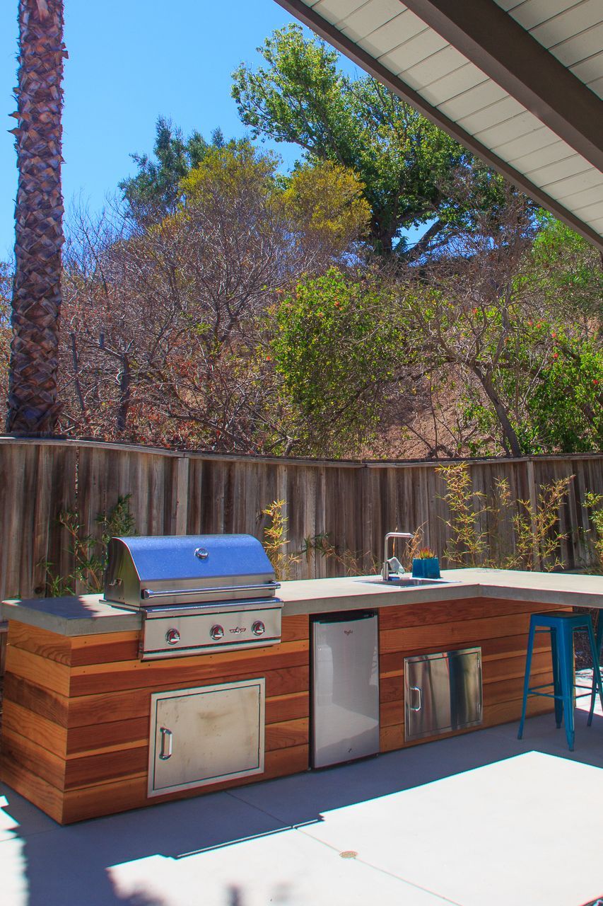 Outdoor kitchen with grill, refrigerator, and countertop, built-in wooden structure. Blue sky.