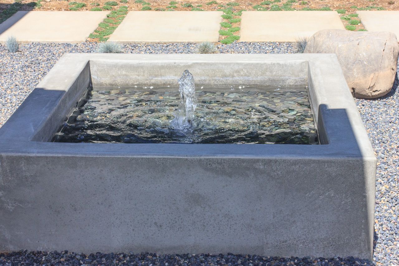 Concrete square fountain with water jet, surrounded by pebbles and a stone slab.