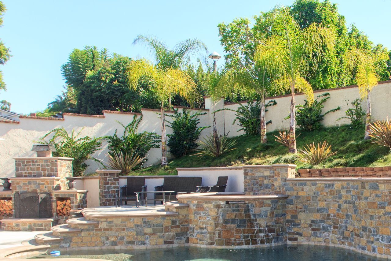 Swimming pool with stone walls, steps, and built-in seating area. Palm trees and greenery on a hillside.