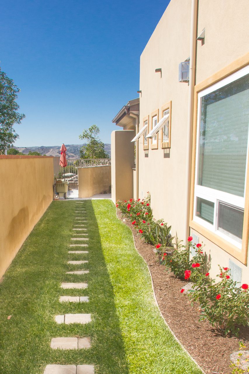 Pathway of stepping stones through green grass beside a tan building with red roses under a blue sky.