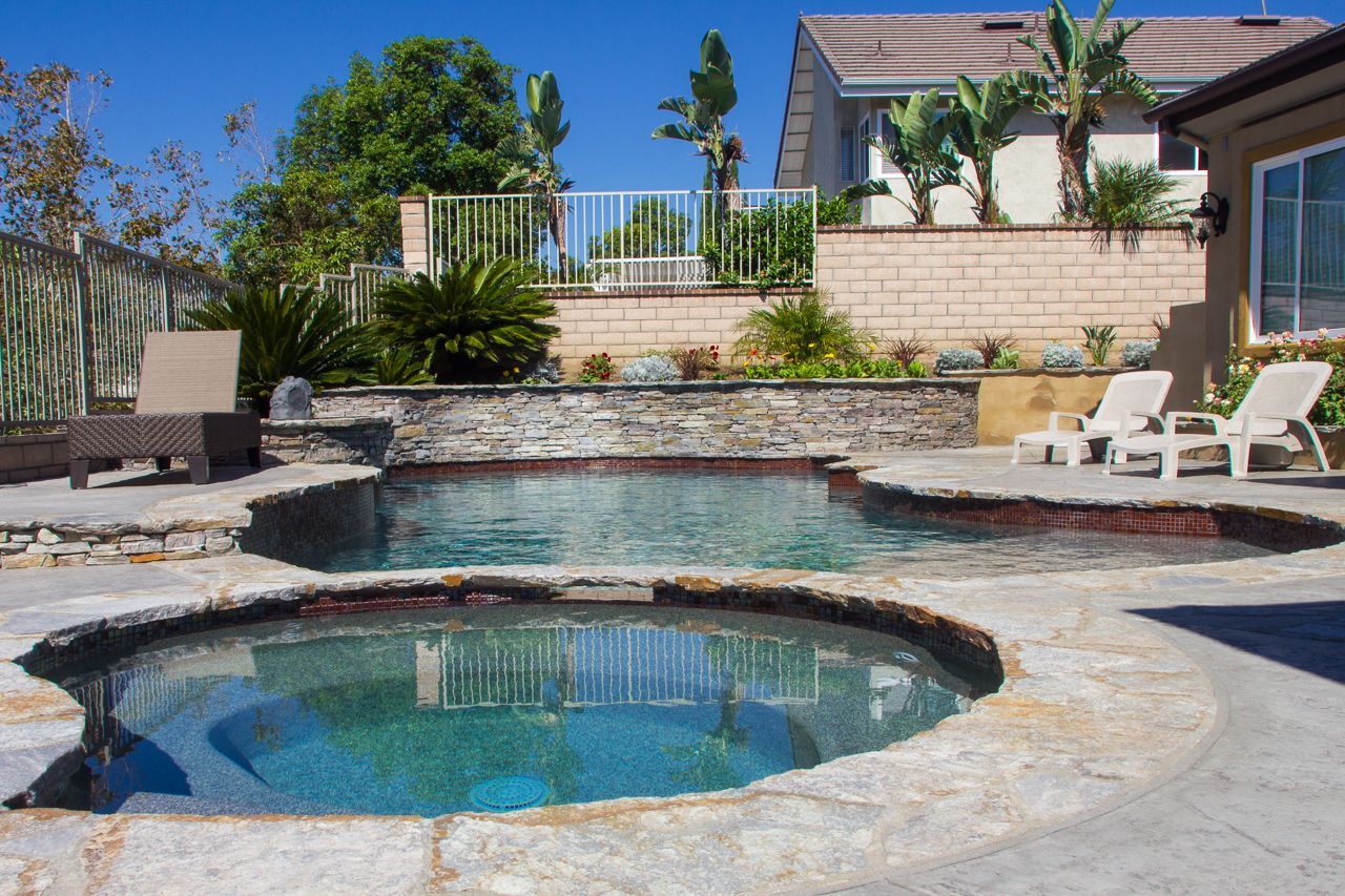 Pool and spa surrounded by stone patio with lounge chairs; lush landscaping and house in the background.