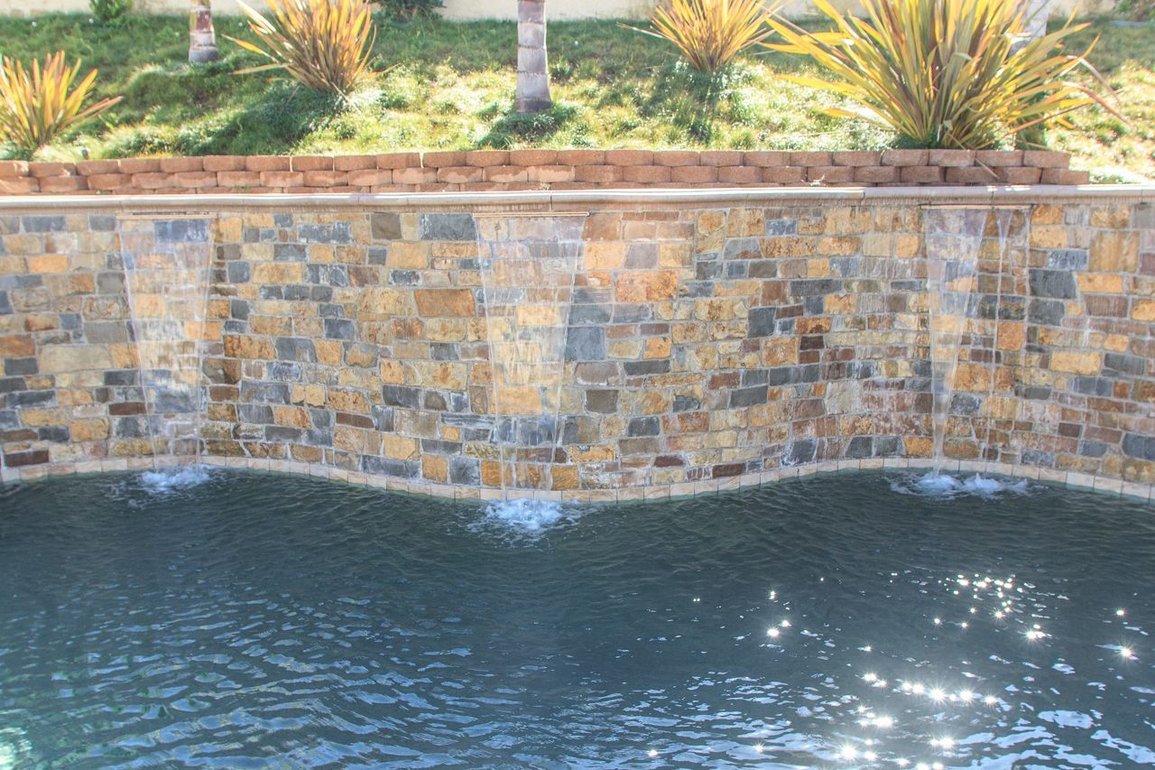 Water cascades from a mosaic tile wall into a pool. Plants and a brick border sit above.