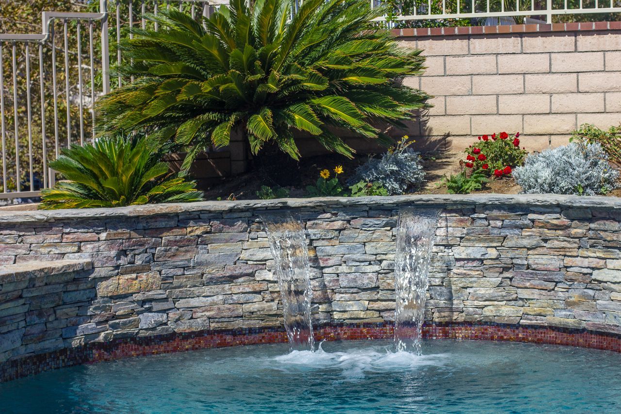 Waterfall feature cascading into a pool, with a stone wall and lush green plants in the background.