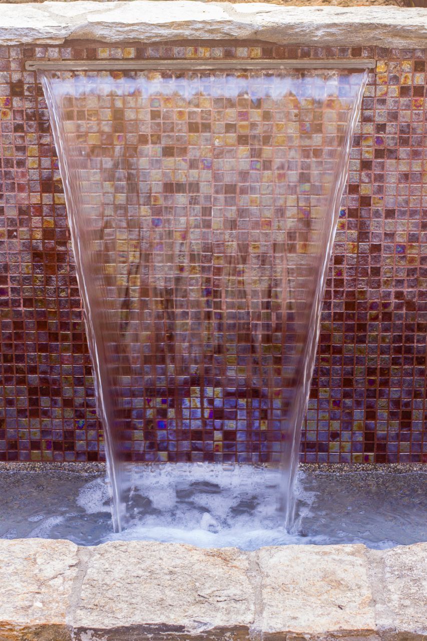 Water cascades down a wide, angled opening against a mosaic tile wall, creating a small pool at the bottom.