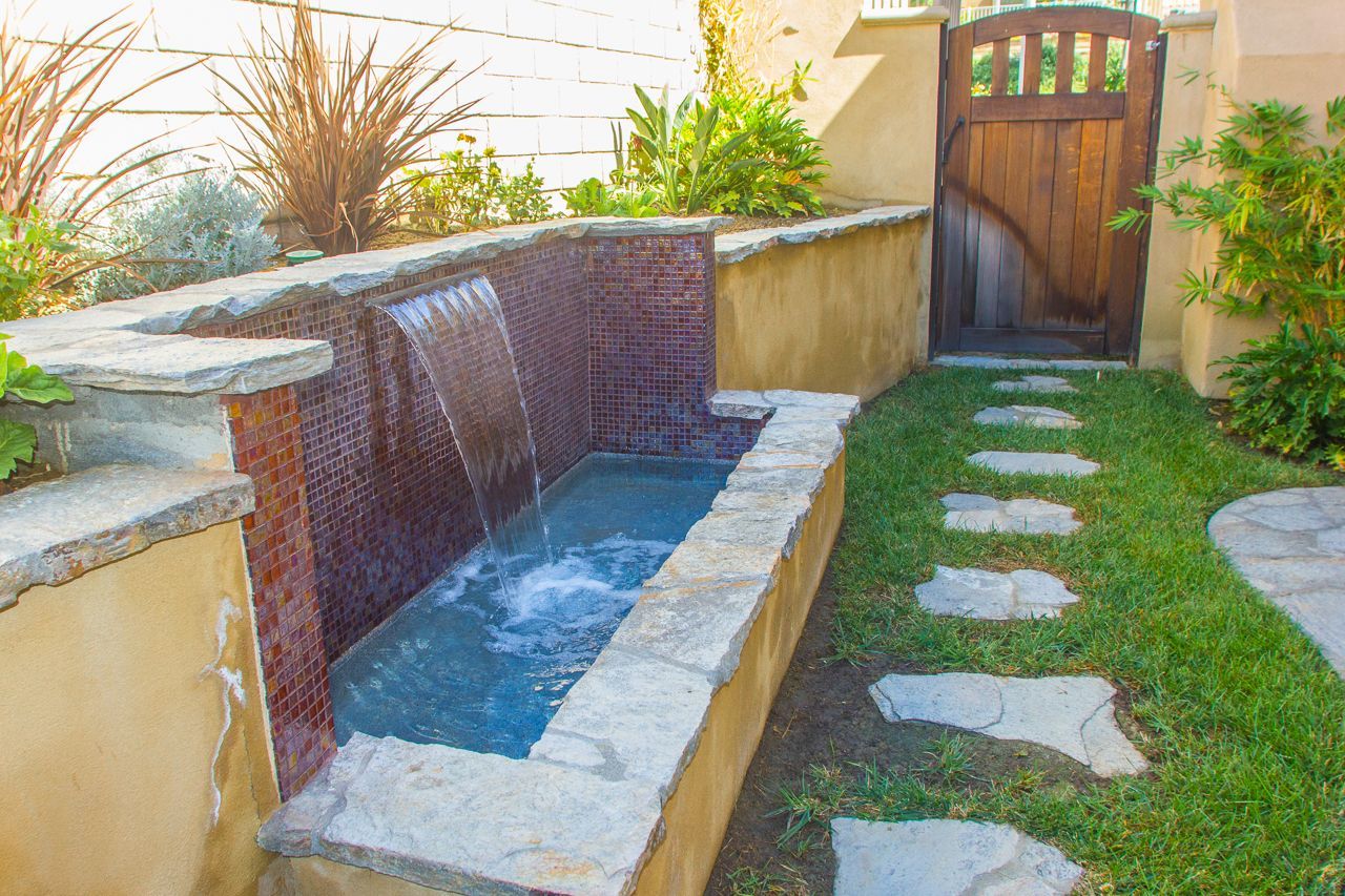 Water feature with a stone basin, cascading water against a tiled wall, a pathway and wooden gate.