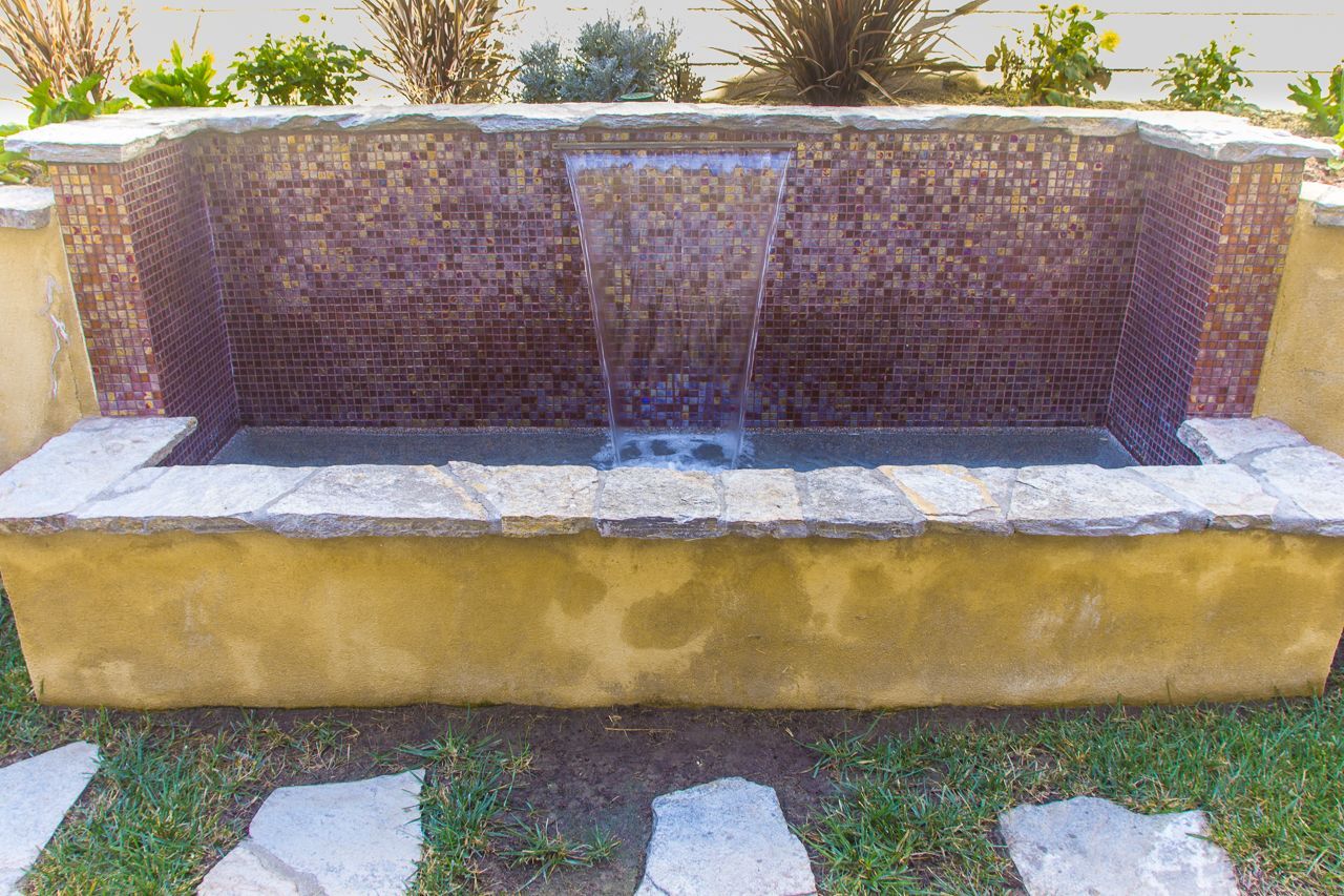 Water fountain with purple tile backdrop and stone accents, flowing water into basin.