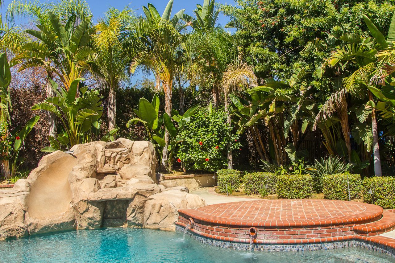 Swimming pool with rock waterfall, palm trees, and a brick-lined hot tub on a sunny day.