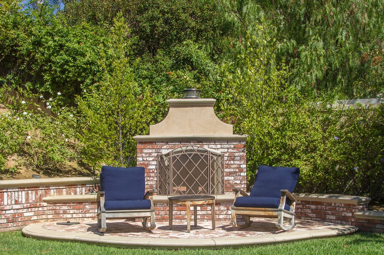 Outdoor brick fireplace with two blue chairs and a small table. Lush greenery in the background.