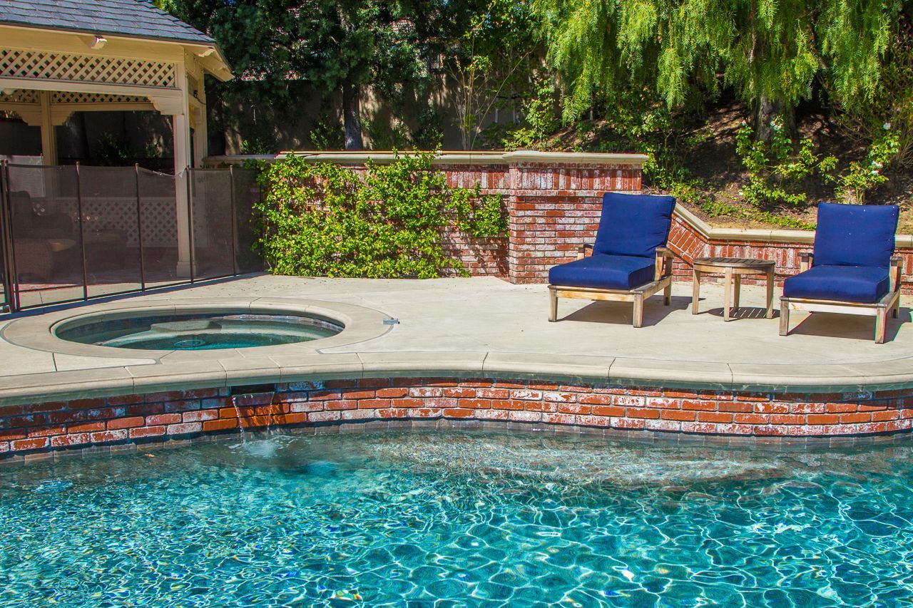 Poolside scene: blue pool and hot tub, two blue lounge chairs, brick wall, gazebo.