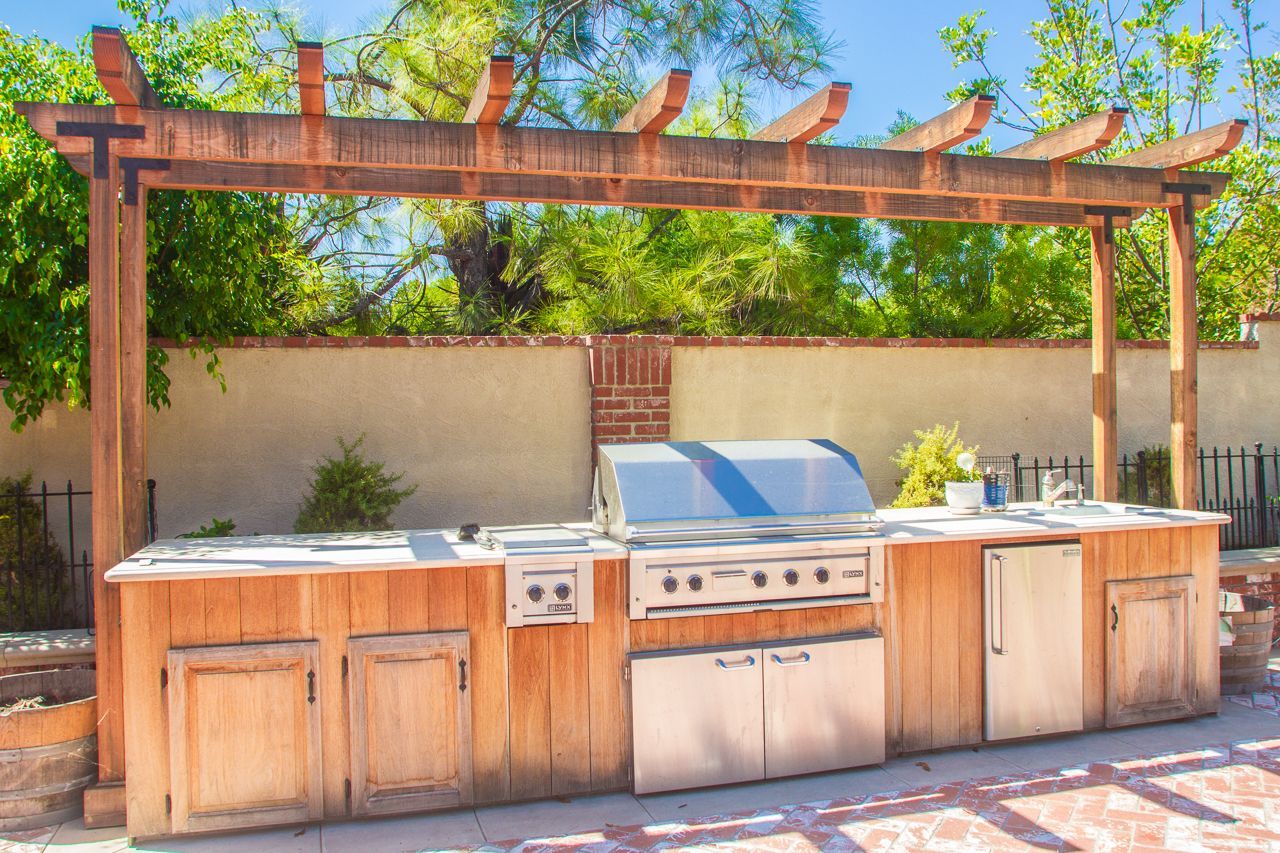 Outdoor kitchen with wooden pergola, stainless steel grill, and refrigerator.