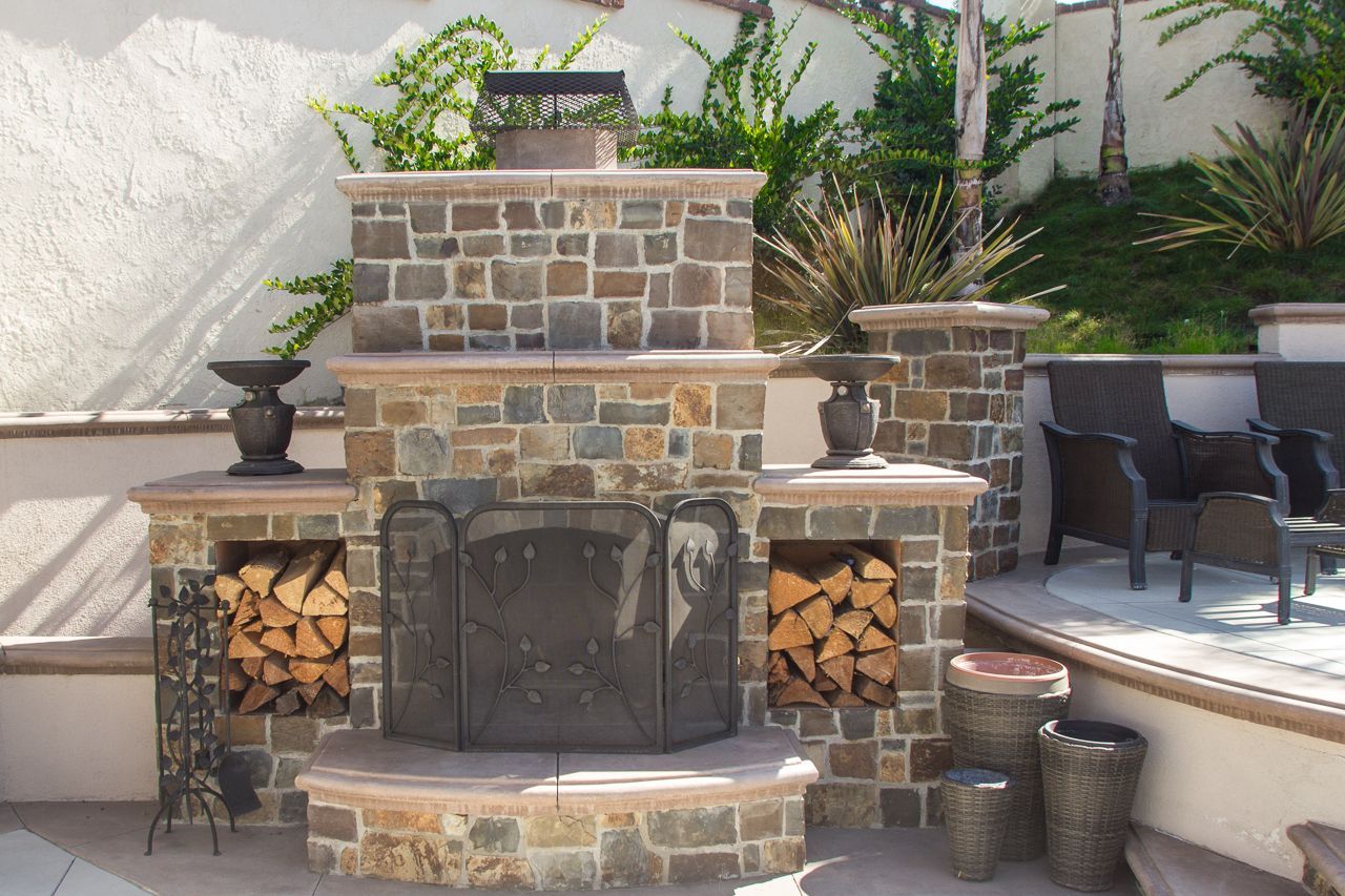 Stone outdoor fireplace with stacked firewood, a metal screen, and decorative urns.