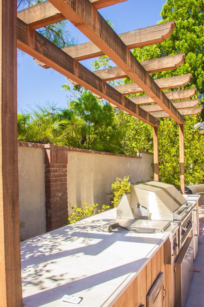 Outdoor kitchen with a wooden pergola, grill, and countertop against a wall.