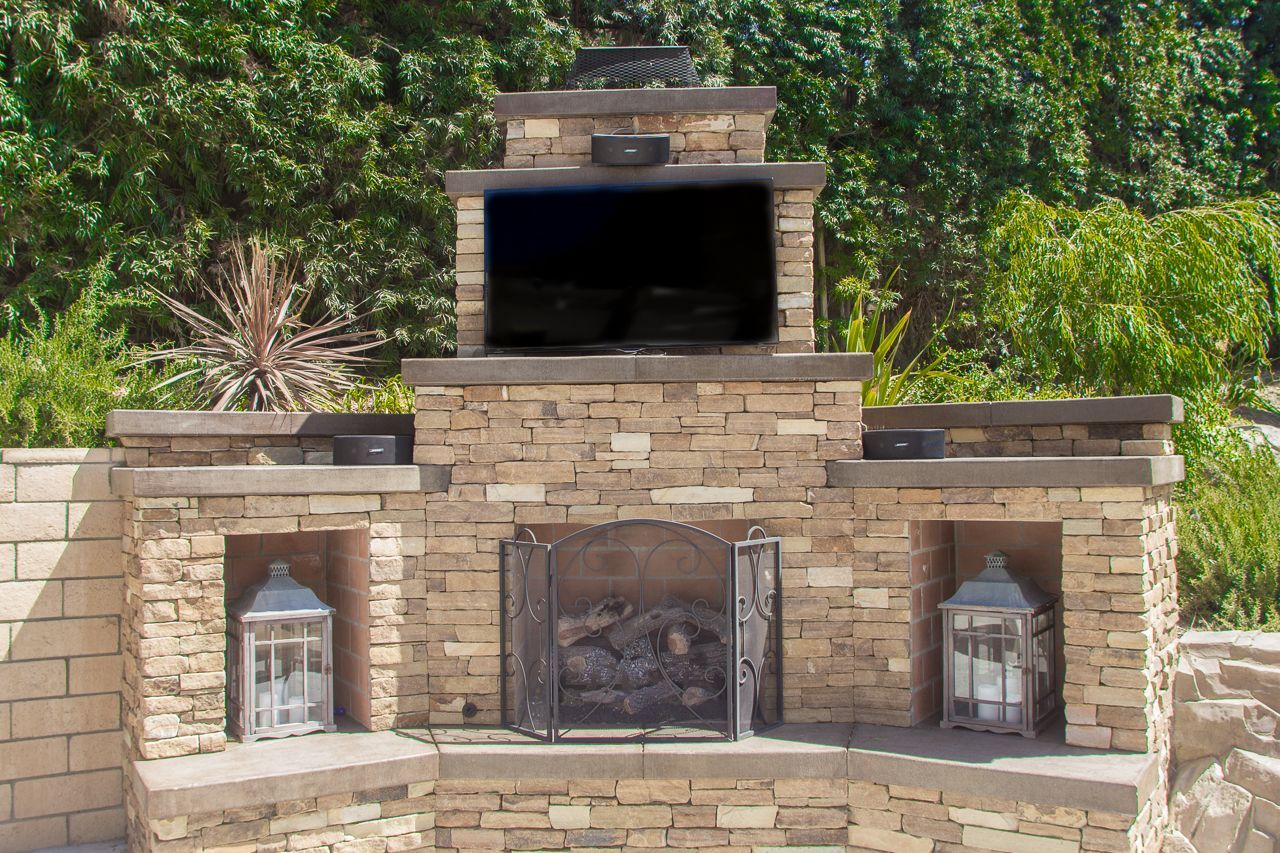 Stone outdoor fireplace with a recessed TV, lanterns, and open firebox.