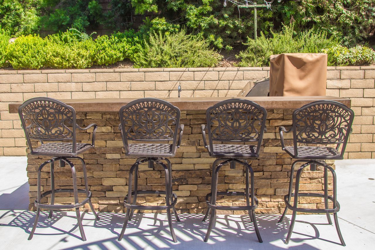 Outdoor bar with four ornate metal swivel bar stools. Brick facade, countertop, and vegetation in the background.