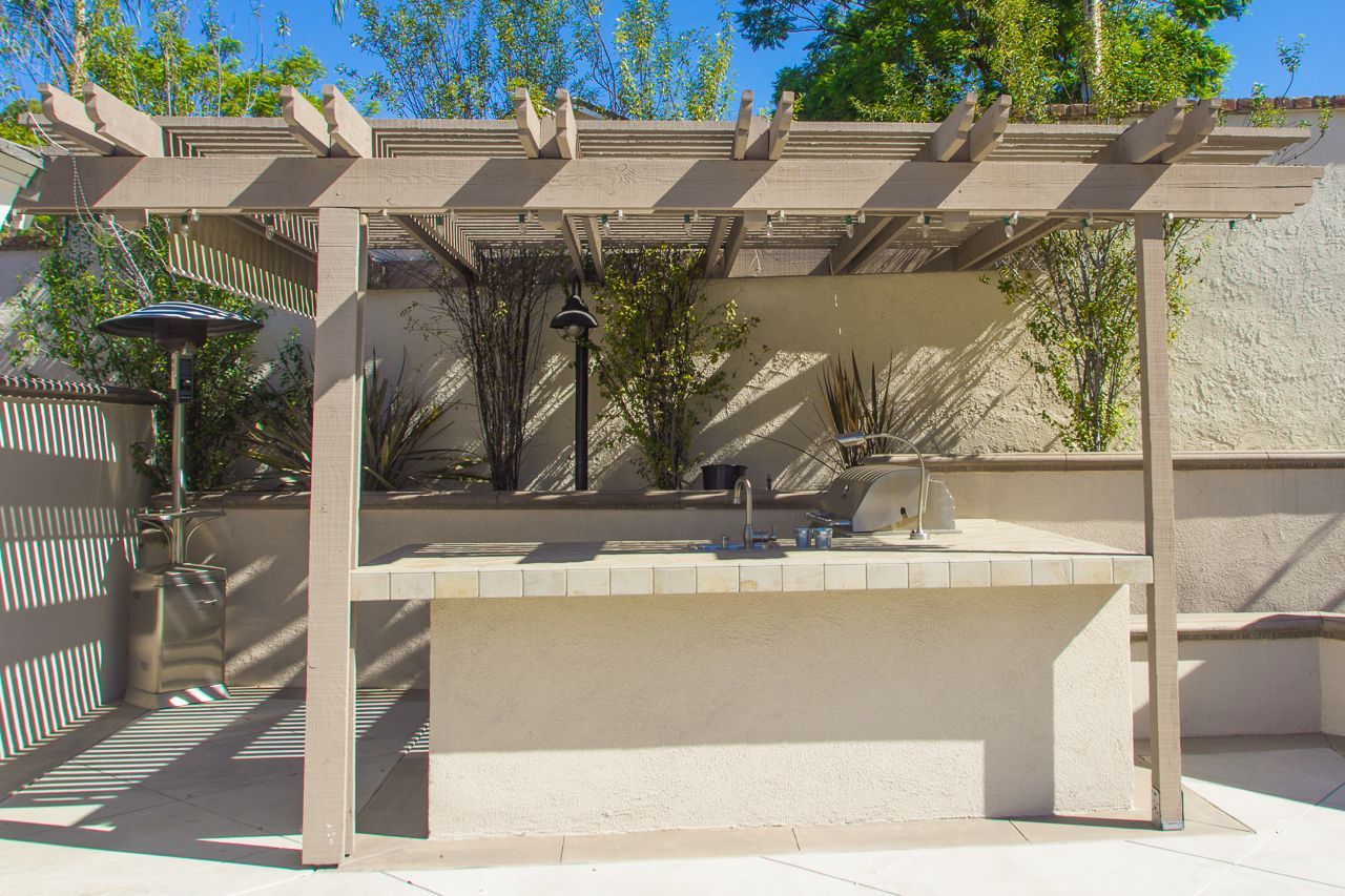 Pergola-covered outdoor kitchen with built-in counter, against a light wall with landscaping, on a sunny day.
