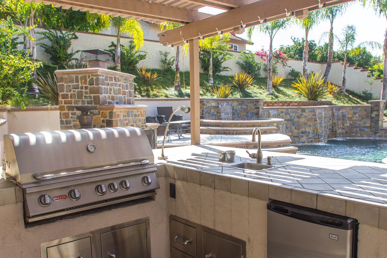 Outdoor kitchen with grill, sink, and stone accents, overlooking a pool and tiered landscape.