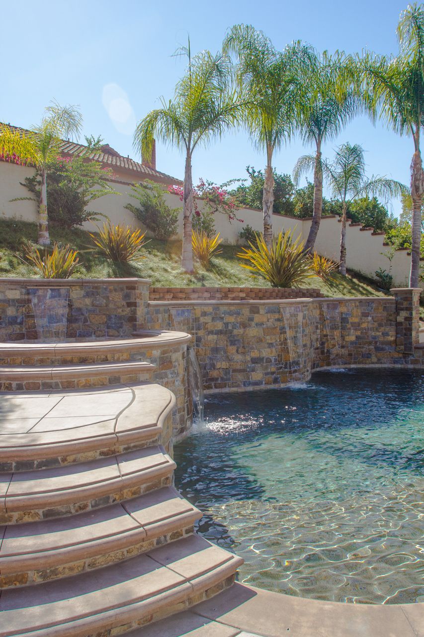 Stone steps lead to a swimming pool with a waterfall feature. Palm trees and hillside in the background under blue sky.