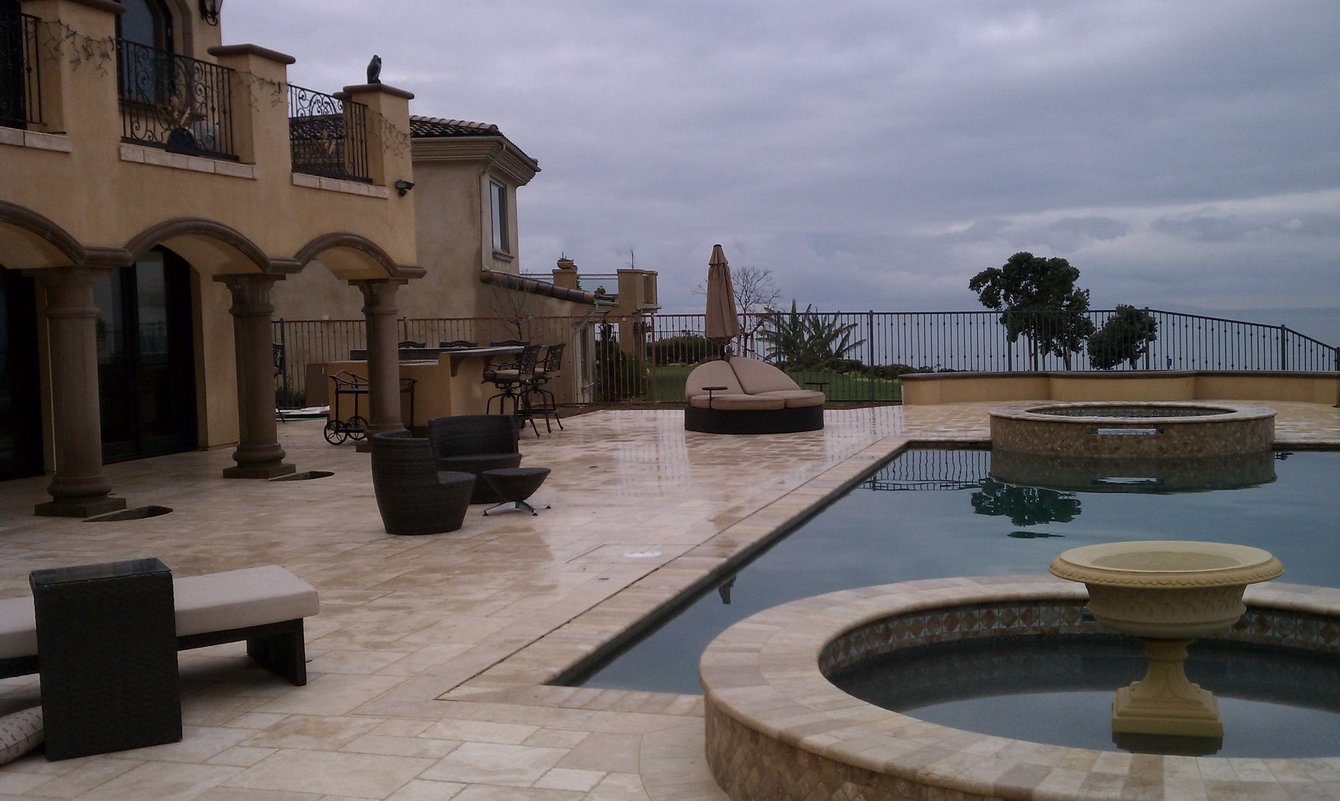 Patio with pool overlooking water, large house with columns, dark clouds.