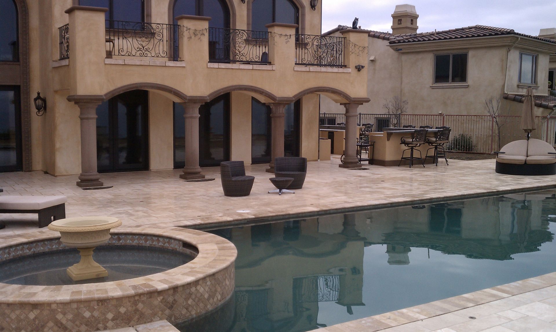 Poolside patio with pool, fountain, and luxury house with arches and columns.