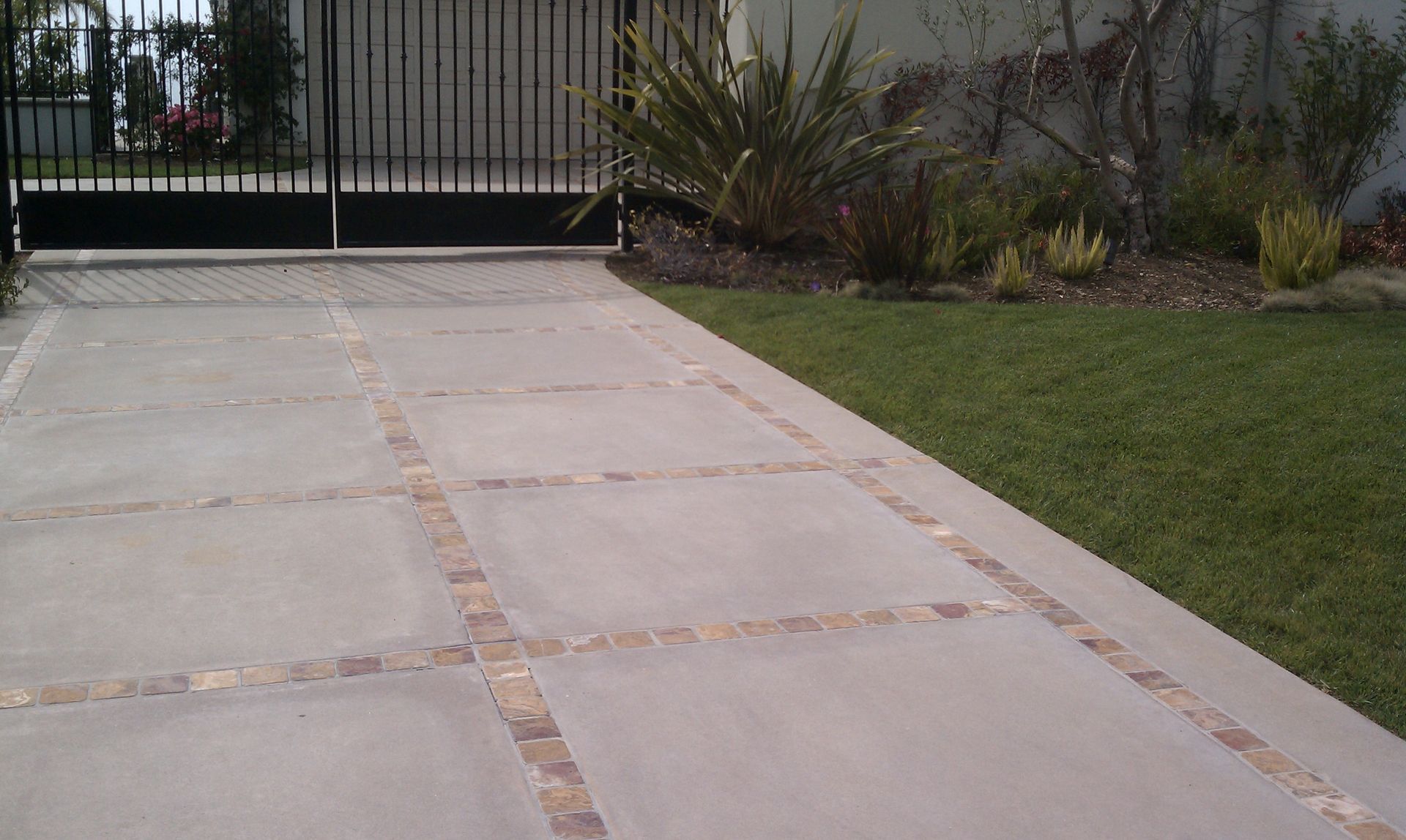 Concrete driveway with brick borders leading to a black gate, beside a green lawn and plants.