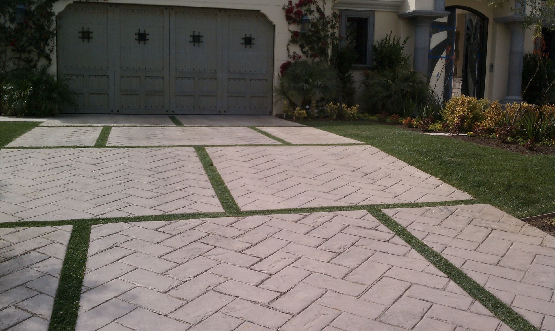 Driveway with beige pavers in herringbone pattern, green grass in between, leading to a gray garage door.