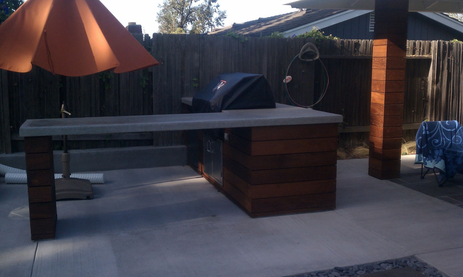 Outdoor kitchen with a concrete countertop, grill, and wooden cabinets under an orange umbrella.