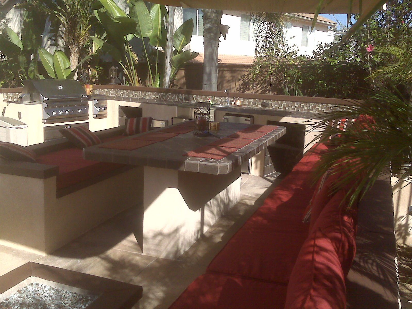 Outdoor kitchen with built-in seating, grill, and table, red cushions, beige structures, and lush greenery in the background.