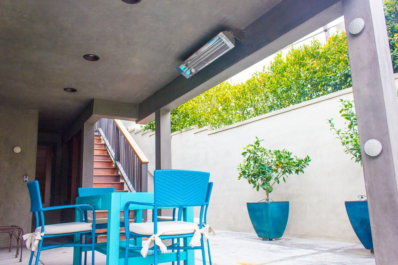 Outdoor patio with blue furniture, a ceiling-mounted heater, and potted plants against a white wall and green hedge.