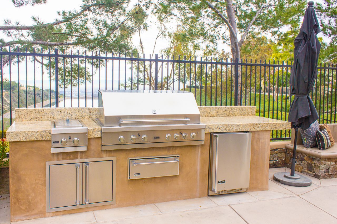 Outdoor kitchen with stainless steel grill, side burner, refrigerator, and storage cabinets.