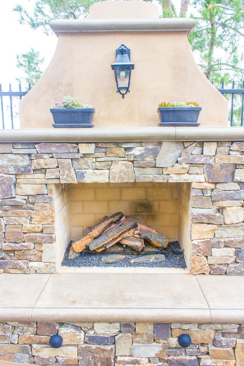 Stone outdoor fireplace with tan stucco top, lantern, and potted plants.