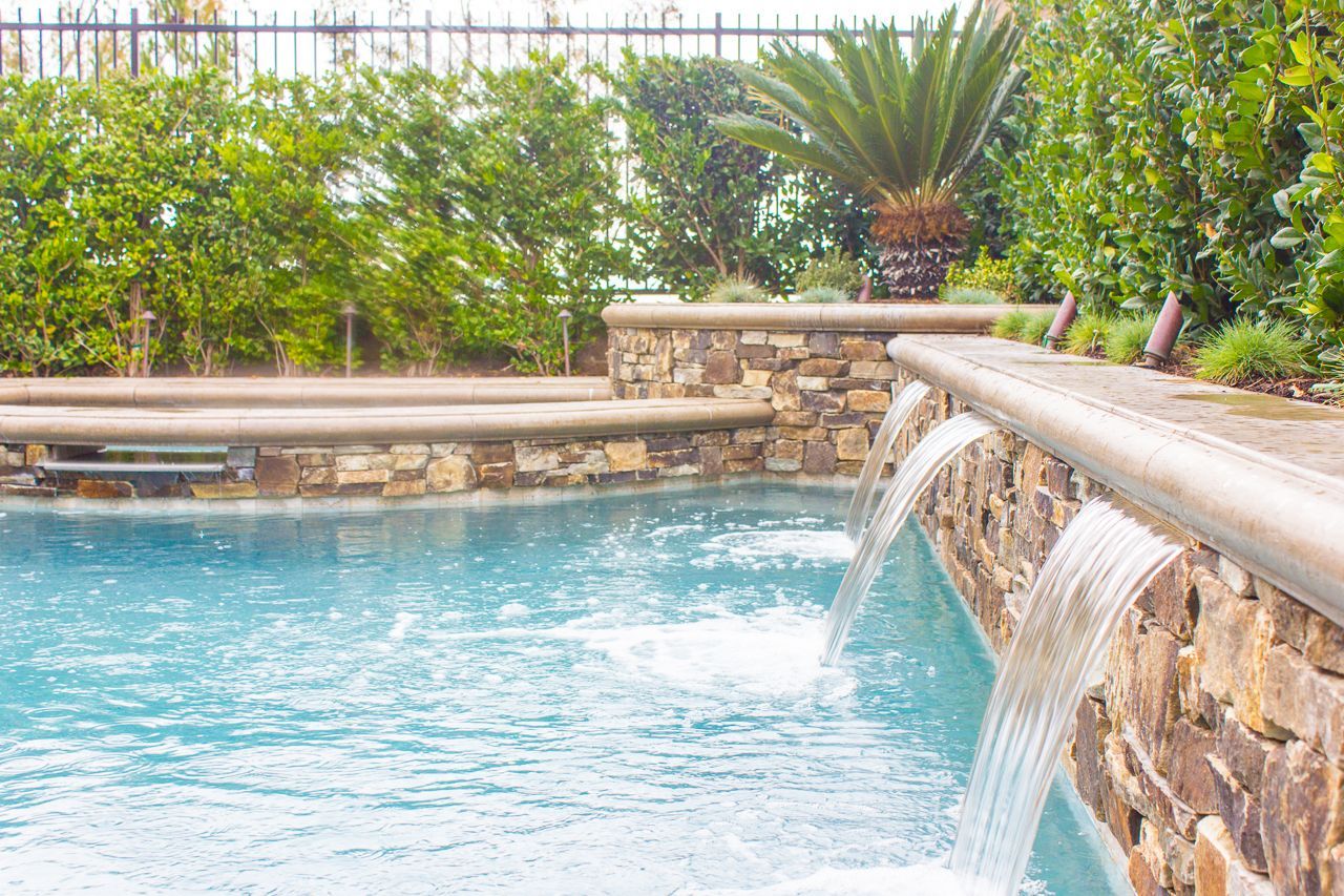 Pool with stone wall and water cascading into the blue water, lush green foliage in background.