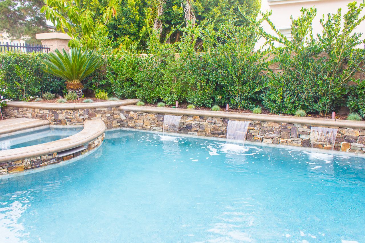 Swimming pool with a stone wall and small waterfalls. Lush green bushes in the background.