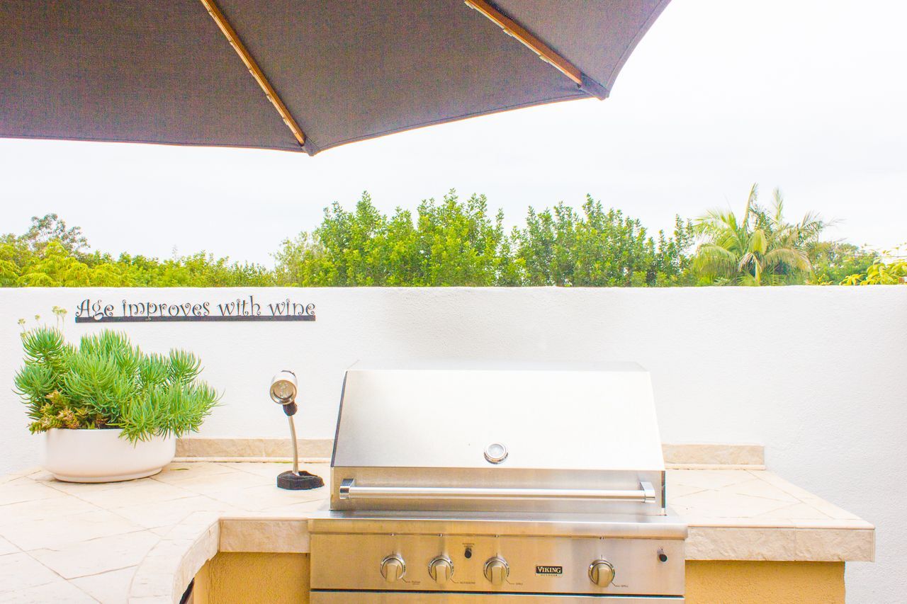 Outdoor stainless steel grill on countertop under a gray umbrella. White wall backdrop with greenery.