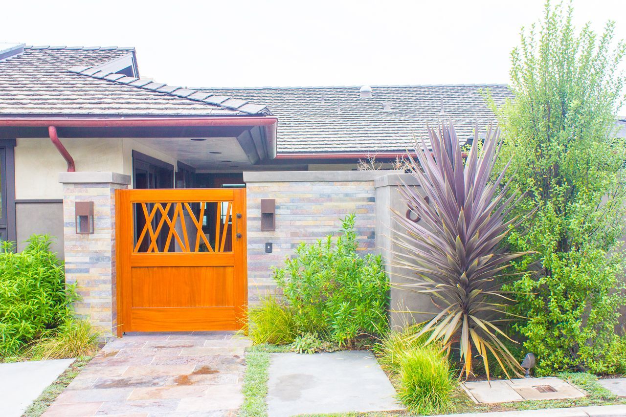 Wooden gate with intricate design, leading to a house, flanked by greenery and stone wall.