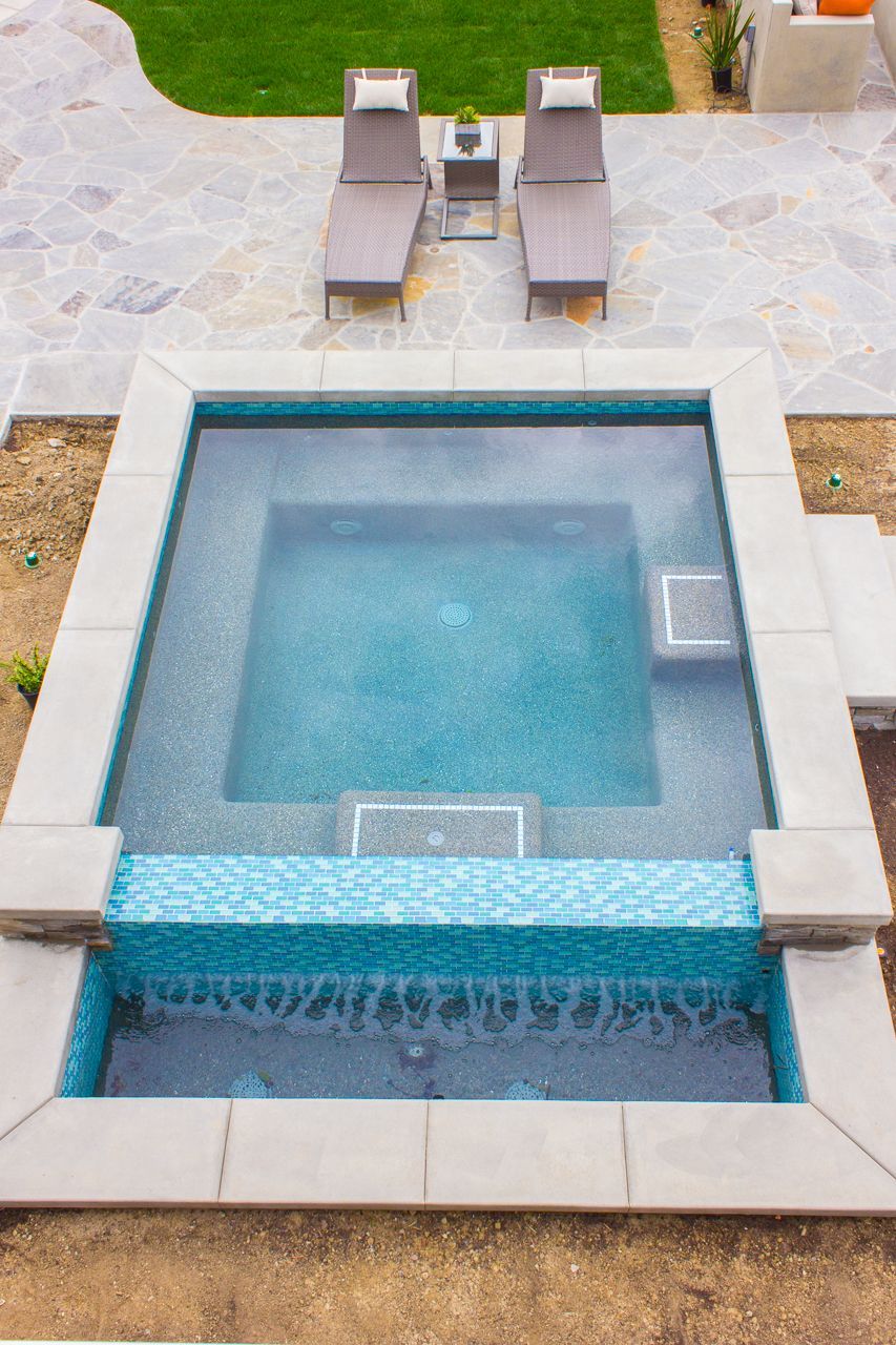 Overhead view of a small square pool with blue mosaic tile and a waterfall feature, surrounded by gray stone and chairs.