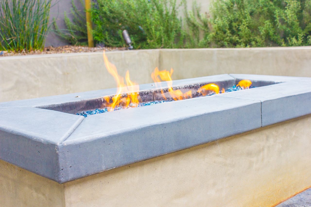 Lit rectangular gas fire pit, surrounded by concrete border, in an outdoor setting.