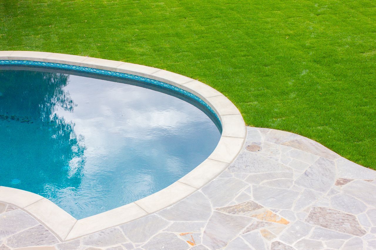 Pool with light stone patio and green lawn reflecting sky.