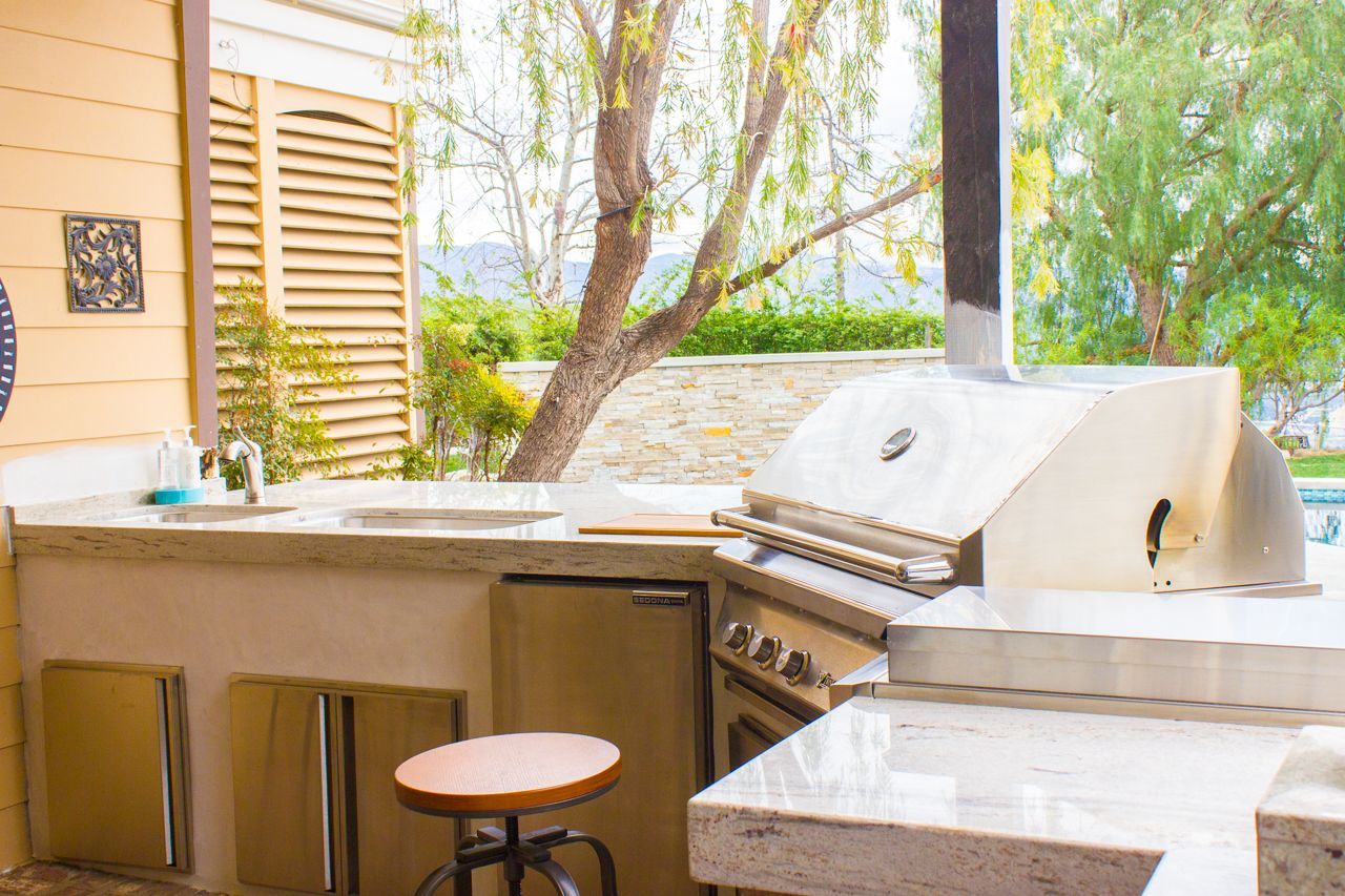 Outdoor kitchen with a stainless steel grill, counter, sink, and a stool.