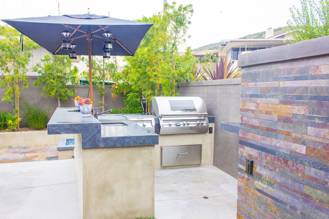 Outdoor kitchen with grill, countertop, and umbrella in a backyard setting.
