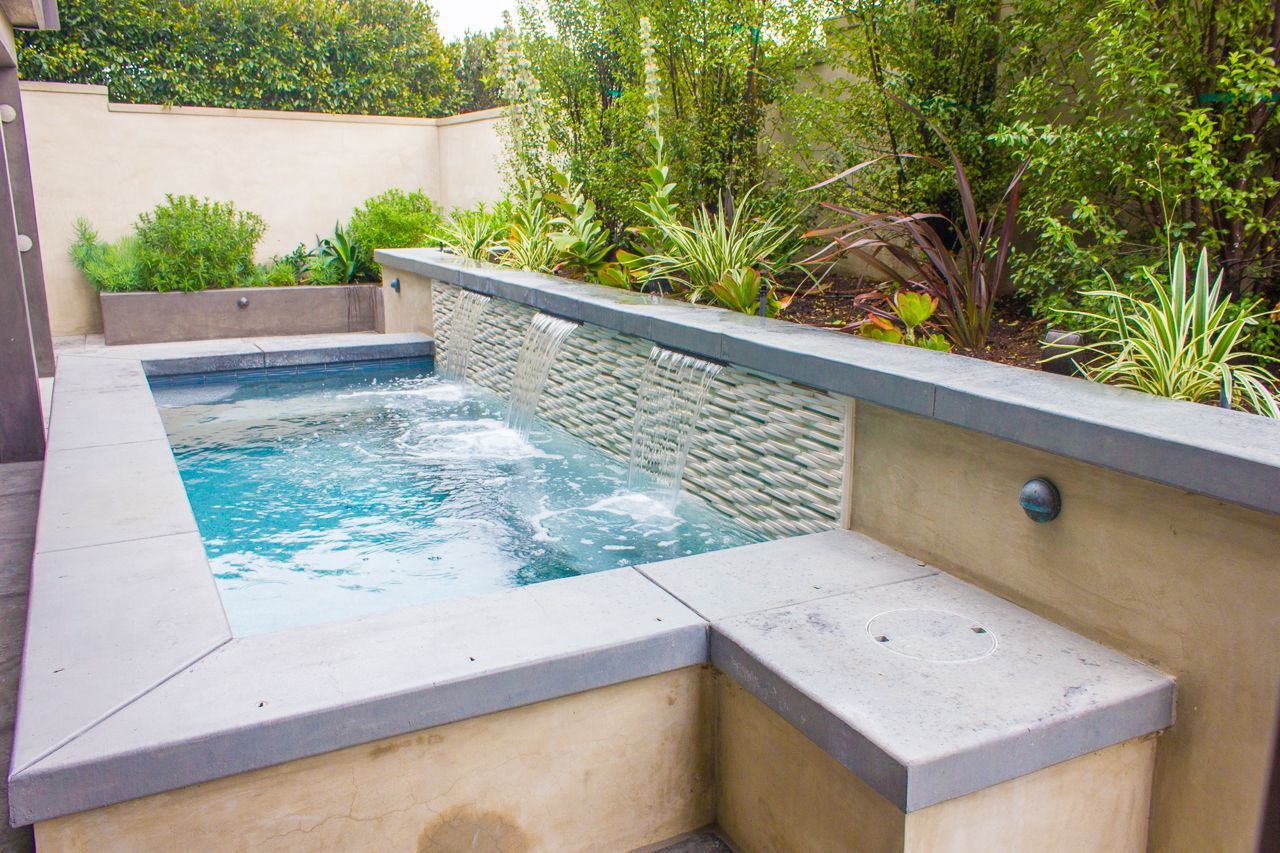 Small backyard pool with waterfall feature and stone wall; plants surround.