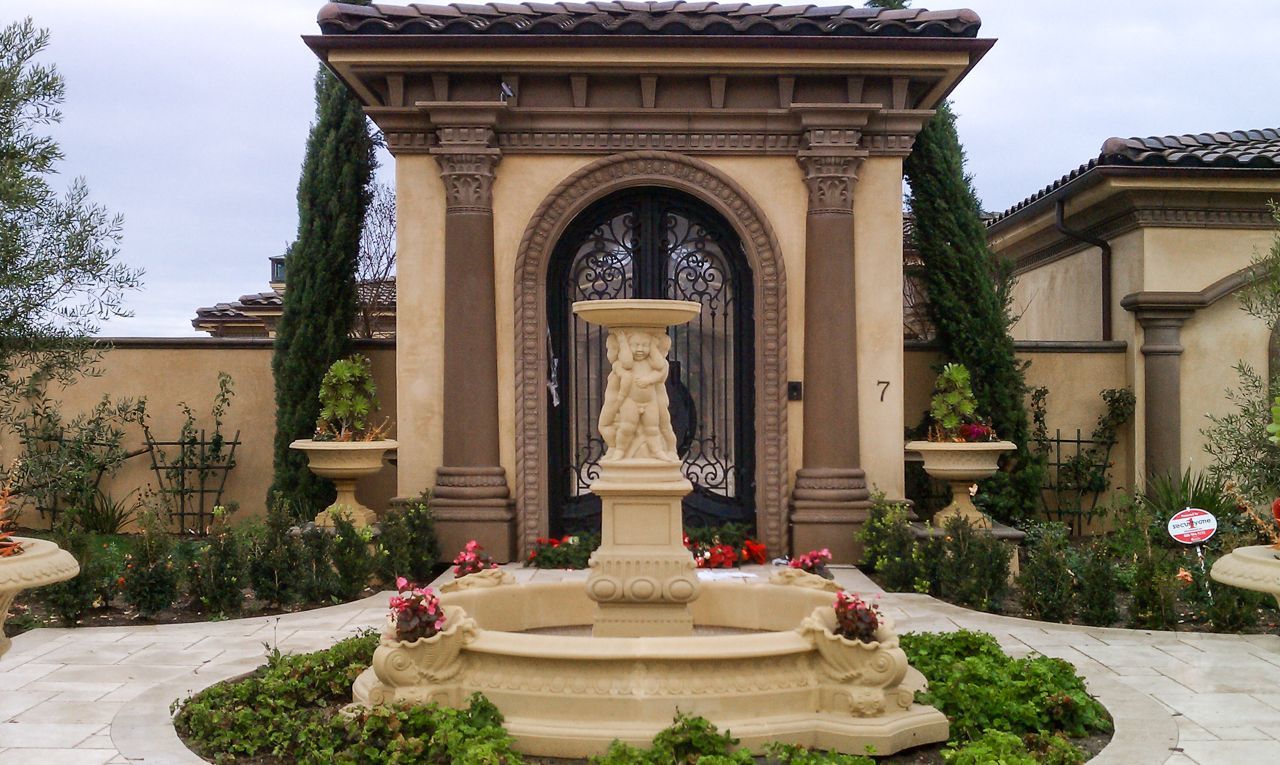 Ornate fountain in a garden setting, with a decorative arched entrance and two flanking pillars.