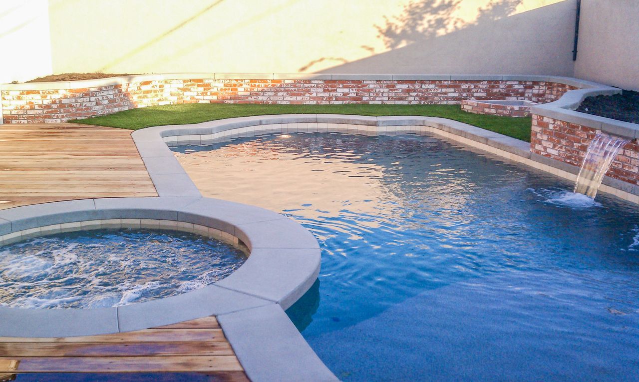 Pool with attached jacuzzi, wooden deck, stone waterfall, and brick retaining wall.