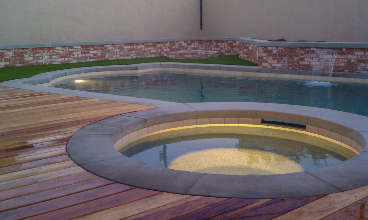 Swimming pool and spa with wooden deck. Water feature and brick wall in background.