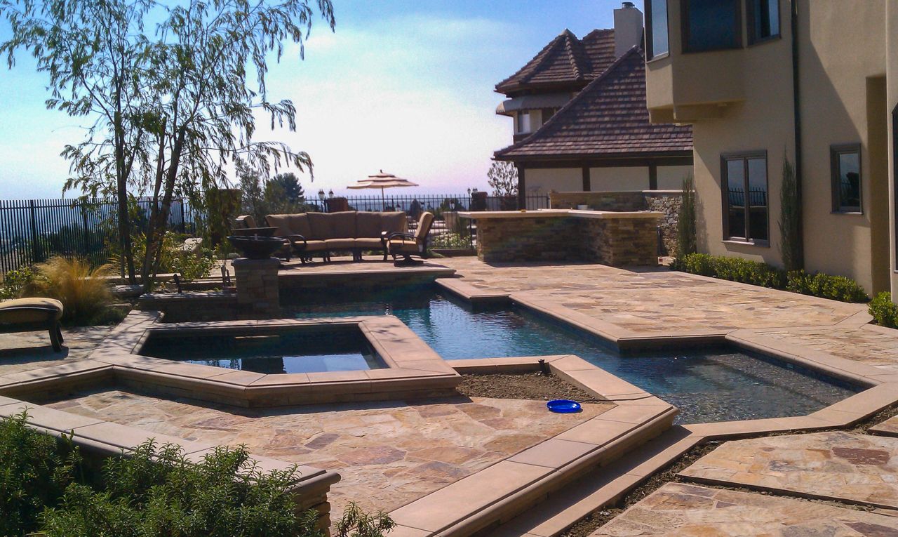 Luxury backyard with a pool, spa, seating area, and ocean view. Stone patio, sunny day.