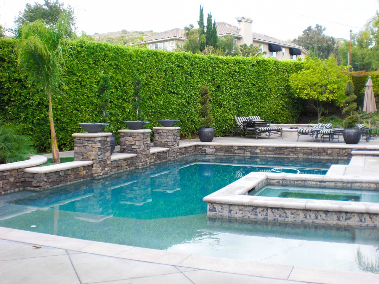 Pool with stone accents, a spa, and a tall green hedge in a backyard setting.