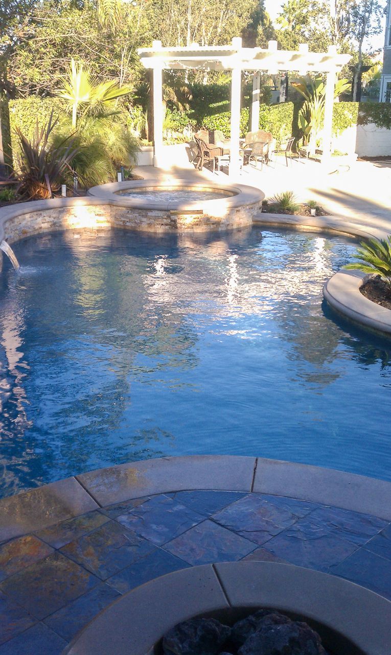 Blue swimming pool with a stone patio, a pergola, and greenery in the background.