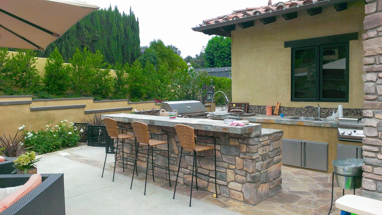 Outdoor kitchen with stone island, seating, grill, and counter, in a patio setting with greenery.